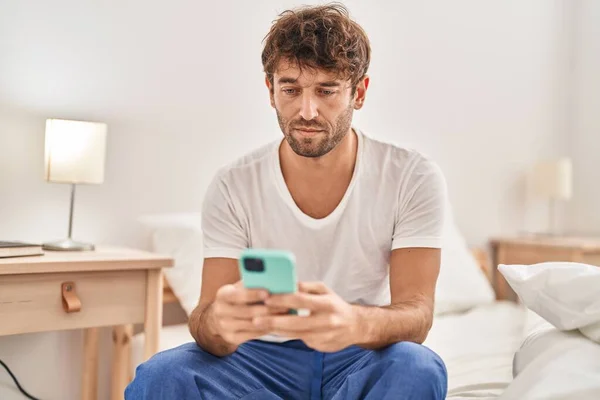 Mladý Muž Používá Smartphone Vážným Výrazem Sedí Posteli Ložnici — Stock fotografie