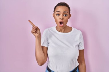 Güzel Afro-Amerikalı kadın pembe arka planda dikilmiş parmağını yana kaldırmış, ağzı açık şaşkın bir ifadeyle şaşırdı.. 