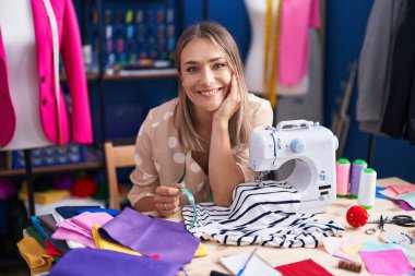 Young caucasian woman tailor smiling confident using sewing machine at sewing studio