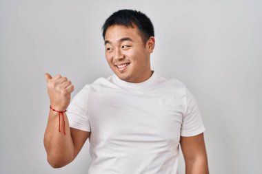 Beyaz arka planda duran genç Çinli adam gülümsüyor, gülümsüyor başparmağıyla yana bakıyor ve işaret ediyor.. 