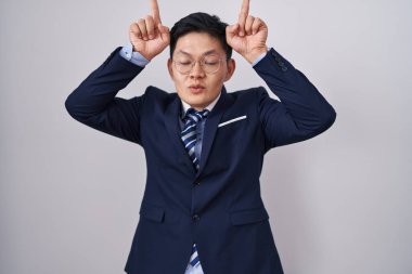 Genç Asyalı takım elbiseli ve kravatlı bir adam parmaklarıyla boynuz gibi komik hareketler yapıyor. 