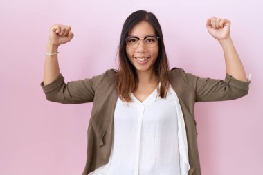 Orta yaşlı Çinli kadın pembe arka planda gözlük takıyor ve gururla gülümseyen kol kaslarını gösteriyor. fitness konsepti. 