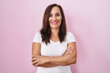 Orta yaşlı esmer kadın pembe arka planda durup kollarını kavuşturup kameraya bakarken gülümsüyor. pozitif kişi. 