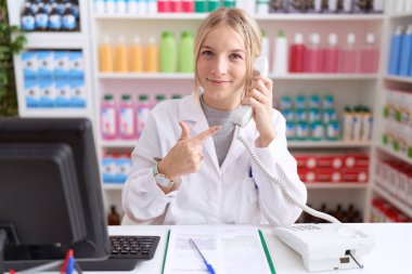 Eczanede çalışan beyaz bir kadın telefonda neşeli bir şekilde konuşuyor. Yüzü gülüyor ve mutlu ve doğal bir ifadeyle işaret ediyor. 