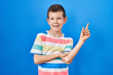 Mavi arka planda duran genç beyaz çocuk yüzünde büyük bir gülümseme ile, kameraya el ile işaret ediyor.. 