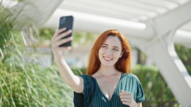 Genç, kızıl saçlı, kendine güvenen bir kadın. Parktaki akıllı telefonun yanında özçekim yapıyor.