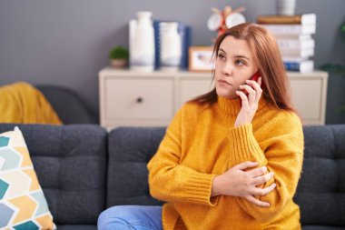 Genç, güzel, iri yarı bir kadın evde endişeli bir ifadeyle akıllı telefondan konuşuyor.