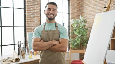 Kollarını kavuşturup sanat stüdyosuna gülümseyen genç Arap sanatçı.