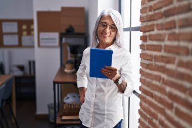 Orta yaşlı, gri saçlı, iş kadını. Ofiste dokunmatik ped kullanarak gülümsüyor.