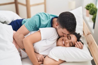 Young hispanic couple lying on bed hugging each other and kissing at bedroom