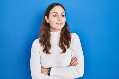 Young hispanic woman standing over blue background looking to the side with arms crossed convinced and confident 
