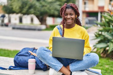African american woman student using laptop sitting on bench at campus park
