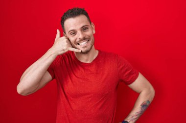Young hispanic man standing over red background smiling doing phone gesture with hand and fingers like talking on the telephone. communicating concepts. 
