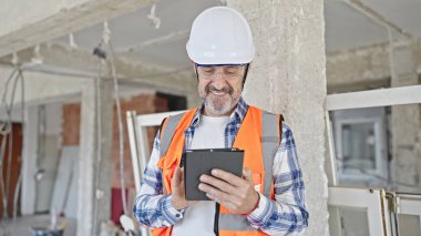 Orta yaşlı inşaatçı inşaat alanında dokunmatik ped kullanarak gülümsüyor.