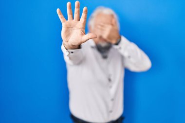 Mavi arka planda gri saçlı orta yaşlı bir adam elleriyle gözlerini kapatıyor ve üzgün ve korku ifadesiyle dur hareketi yapıyor. utangaç ve olumsuz kavram. 