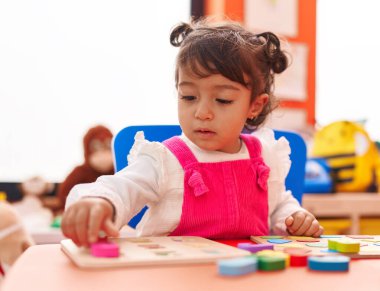 Adorable hispanic girl playing with maths puzzle game sitting on table at kindergarten