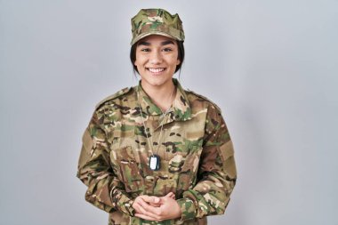 Güney Asyalı genç bir kadın kamuflaj üniforması giyiyor ve elleri birarada ve mutlu ve rahat bir şekilde gülümsüyor. Başarı ve iyimserlik 