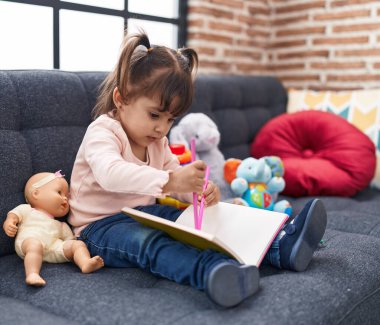 Adorable hispanic girl drawing on notebook sitting on sofa at home