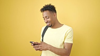 Sırt çantası takan Afrikalı Amerikalı bir adam izole edilmiş sarı arka plan üzerinde akıllı telefon kullanıyor.