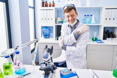 Laboratuvarda çalışan beyaz bir adam mutlu ve olumlu bir şekilde kendine sarılıyor, kendinden emin gülümsüyor. Sevgi ve özgüven. 