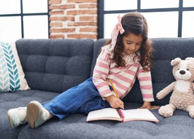 Adorable hispanic girl drawing on notebook sitting on sofa at home
