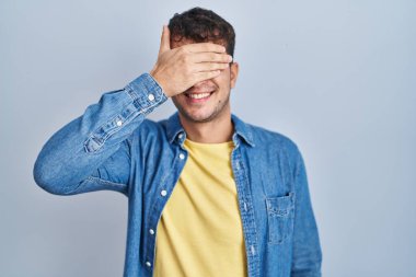 Mavi arka planda dikilen genç İspanyol adam. Gülümsüyor ve gülümsüyor. Gözlerini şaşkınlıkla kapatıyor. kör kavram. 