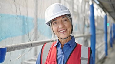 Orta yaşlı, gri saçlı kadın müteahhit sokakta kendine güvenerek gülümsüyor.