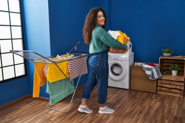 Genç, güzel, İspanyol bir kadın. Çamaşır odasında elinde sepetle gülümsüyor.