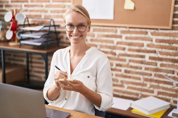 Young blonde woman business worker using laptop writing on reminder paper at office