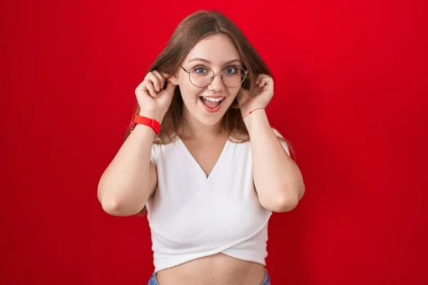 Young Caucasian Woman Standing Red Background Smiling Pulling Ears Fingers — Stock Photo, Image