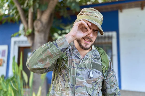 Young hispanic man wearing camouflage army uniform outdoors smiling happy doing ok sign with hand on eye looking through fingers