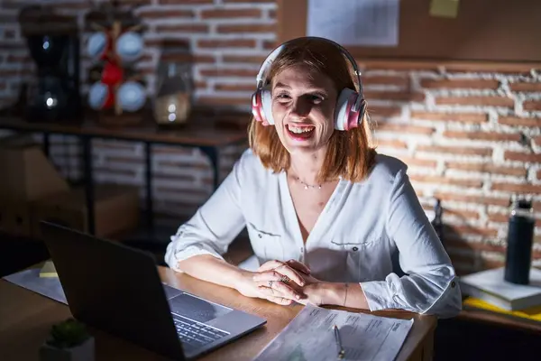 Young redhead woman working at the office at night wearing headphones smiling and laughing hard out loud because funny crazy joke.