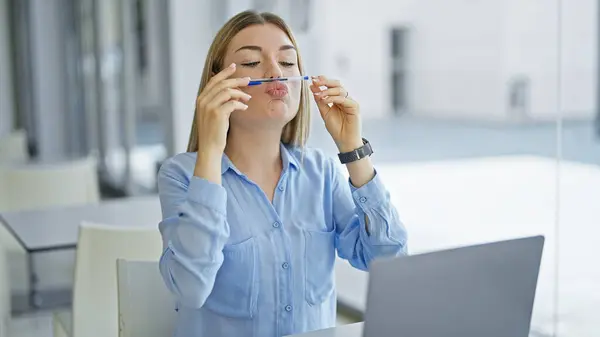 Young blonde woman business worker with pen between nose and mouth doing funny gesture at office