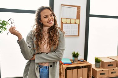 Young beautiful hispanic woman business worker standing with arms crossed gesture holding glasses at office