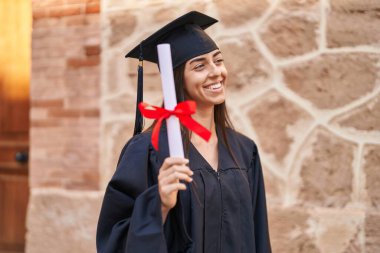 Young hispanic woman wearing graduated uniform holding diploma at university