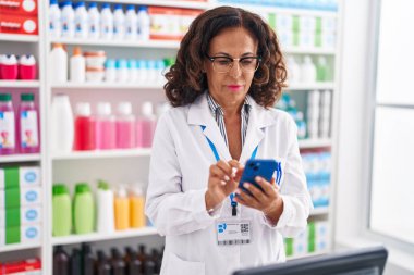 Middle age woman pharmacist using smartphone working at pharmacy