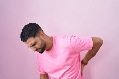 İspanyol genç adam pembe arka planda duruyor sırt ağrısı çekiyor, elleriyle dokunuyor, kas ağrısı çekiyor. 