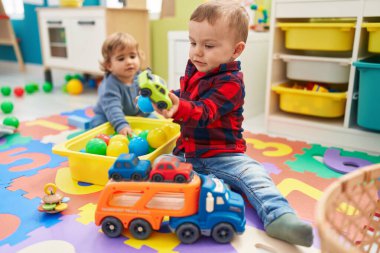 Anaokulunda yerde oturan top ve araba oyuncaklarıyla oynayan sevimli çocuklar.