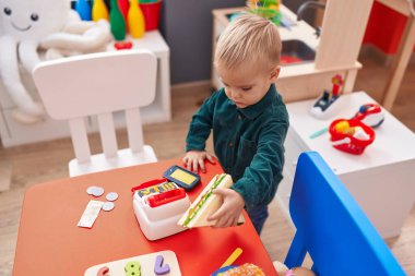 Anaokulunda elinde sandviç ve süpermarket oyuncağı tutan sevimli beyaz çocuk.