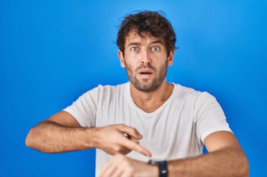 Hispanic young man standing over blue background in hurry pointing to watch time, impatience, upset and angry for deadline delay 
