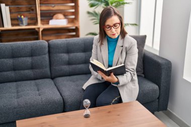 Orta yaşlı kadın psikolog psikoloji merkezindeki koltukta oturmuş kitap okuyor.