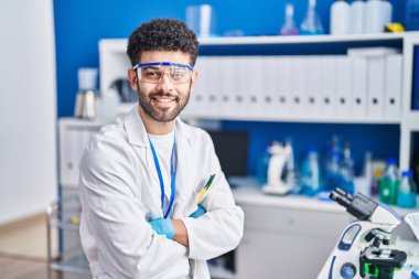 Bilim adamı üniforması giyen genç bir Arap laboratuvarda kollarını kavuşturup oturuyor.