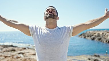 Genç İspanyol adam kumsalda gözleri kapalı ve kolları açık nefes alıyor.