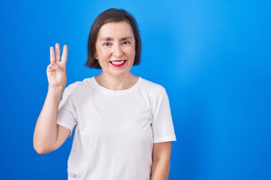 Middle age hispanic woman standing over blue background showing and pointing up with fingers number three while smiling confident and happy. 