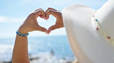 Genç, güzel Latin kadın turist. Yazlık şapka takıyor. Plajda kalp sembolü yapıyor.