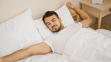 Young hispanic man waking up stretching arms at bedroom