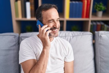 Orta yaşlı bir adam akıllı telefonda konuşuyor. Evdeki kanepede oturuyor.