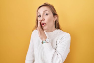 Beyaz süveter giyen genç beyaz bir kadın. Sarı arka plan. Ağız üstüne el. 