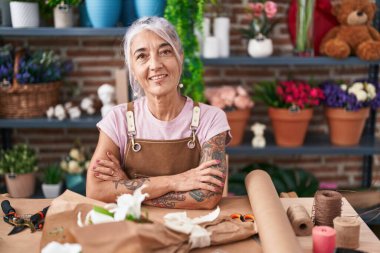 Orta yaşlı, kır saçlı, çiçekçiye kollarını kavuşturup gülümseyen bir çiçekçi kadın.