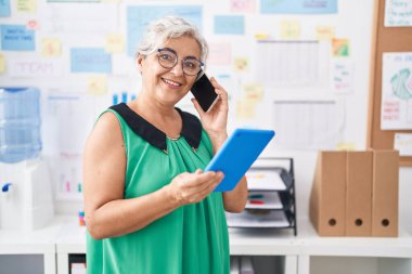 Orta yaşlı, kır saçlı, iş kadını. Ofiste dokunmatik ped kullanarak akıllı telefondan konuşuyor.